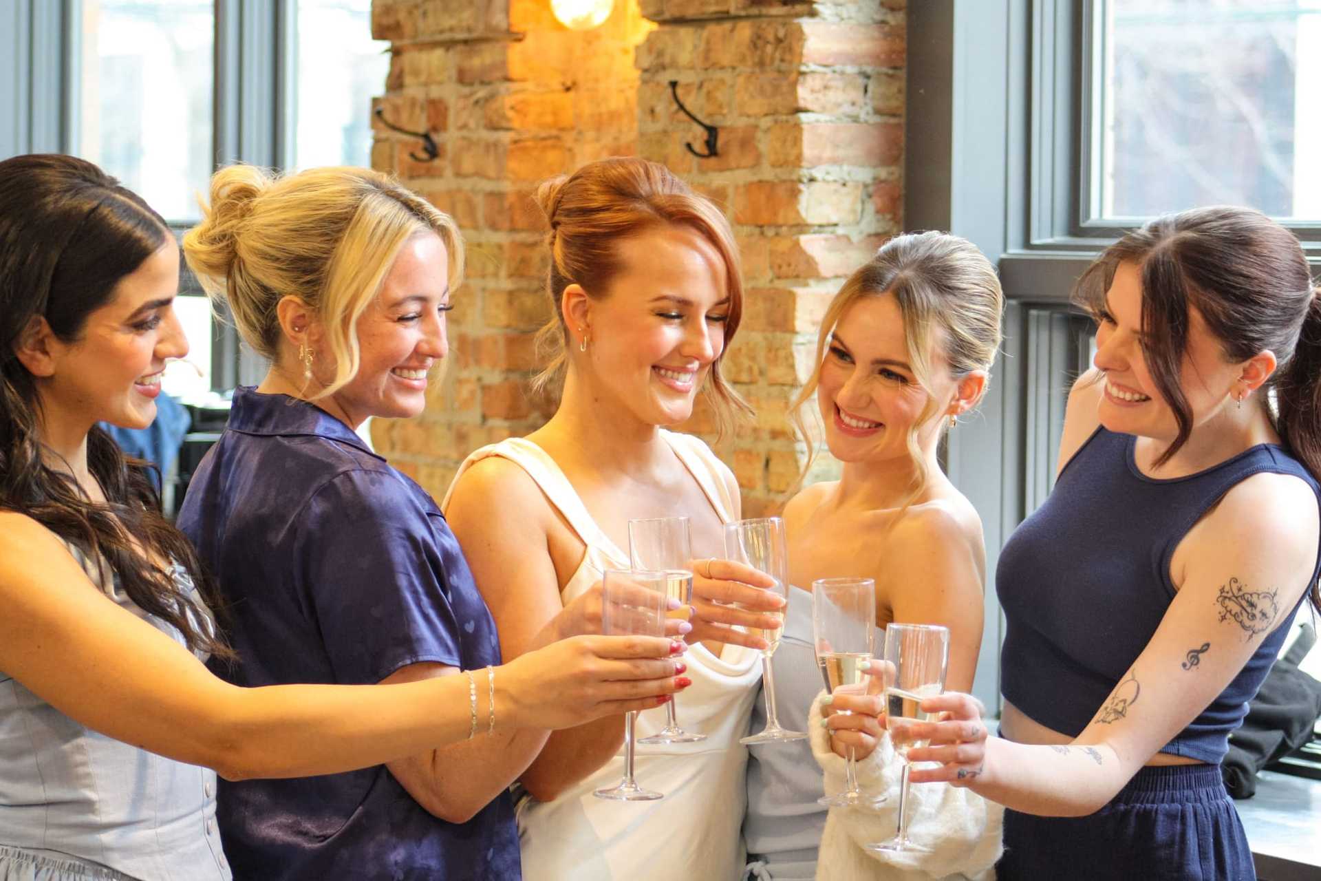 Bridesmaids and bride smiling and toasting with champagne glasses indoors.