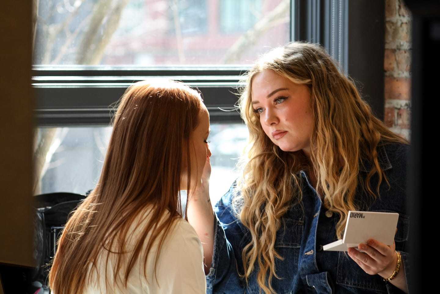 Two women indoors; one applying makeup to the other's face near a window.